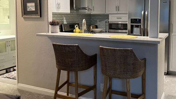 A modern kitchen with grey cabinets, a stainless steel refrigerator, a microwave, and a coffee maker. There are two wicker bar stools and a small island with a yellow placemat. - Prestigous Location within LBUSD. Mid to Long Term Discounts. Views of Foothills (Aliso Viejo)