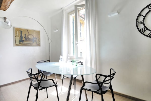 A dining area with a round glass table, black chairs, a painting on the wall, and a large window with curtains.