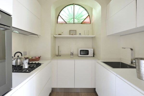 Una cocina compacta con armarios blancos, estufa a gas, microondas y una ventana con lámpara de vidrio verde.
