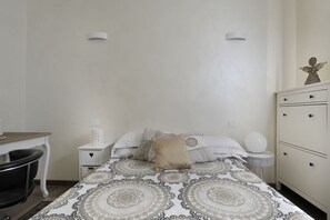 A bedroom with a patterned bedspread, a bedside table with a lamp, a dresser, and a small round table with a lamp.