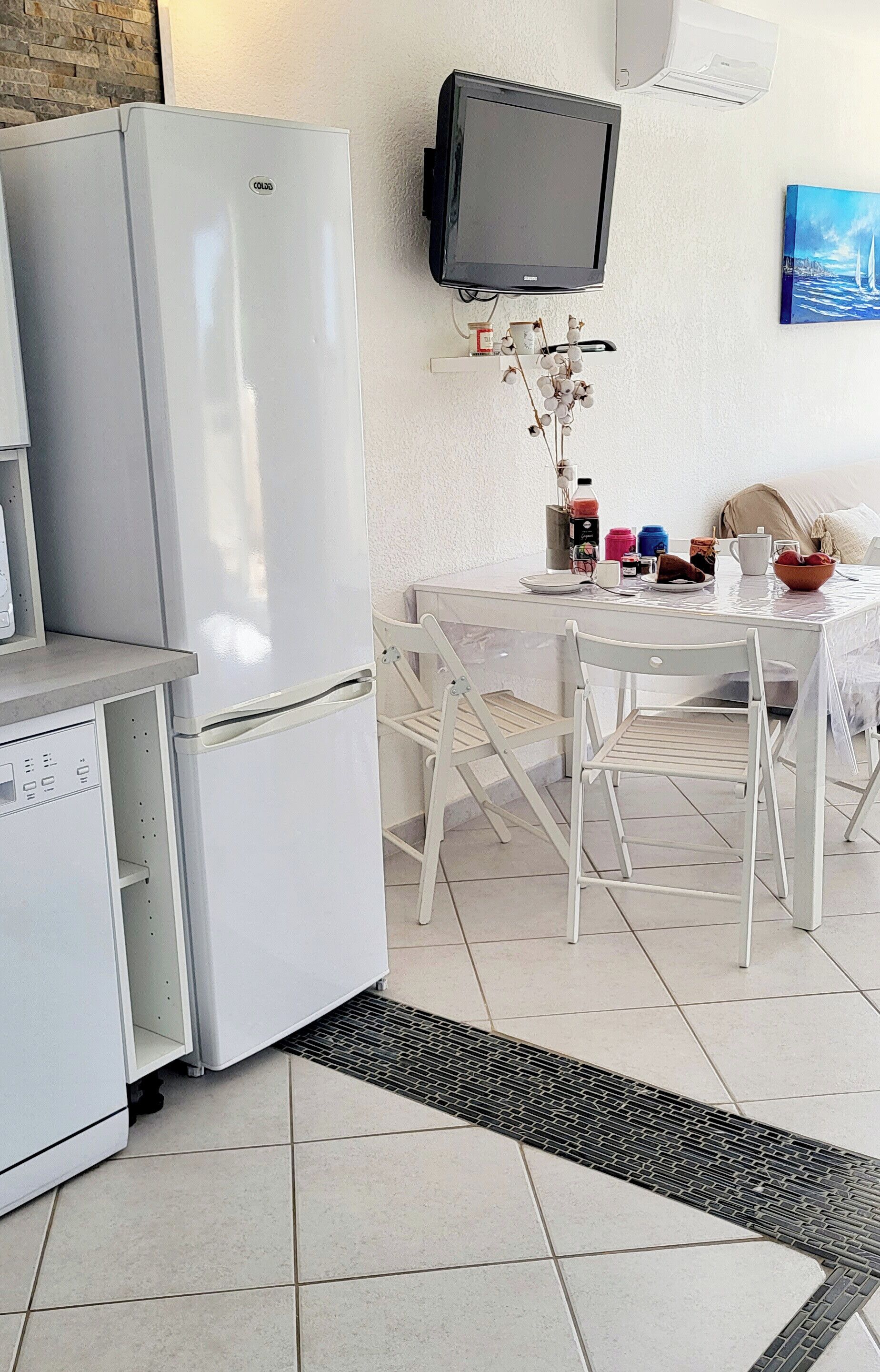 A compact kitchen with a white refrigerator, a small dishwasher, a dining table with chairs, a mounted television, and a tiled floor.