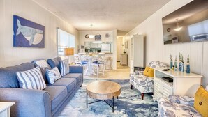 A living room with a sofa, coffee table, and patterned ottoman. There is a dining area with a table and chairs in the background. - Beach Condo. Across from Ocean Annie's. 1st floor beach condo with large pool. (Myrtle Beach)