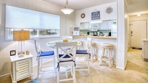 A compact kitchen with white cabinetry, a dining area with white chairs, a small bar stool, and a hanging light fixture. - Beach Condo. Across from Ocean Annie's. 1st floor beach condo with large pool. (Myrtle Beach)