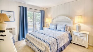 A bedroom with a bed, two bedside tables, a lamp, a picture frame, and a window with curtains. - Beach Condo. Across from Ocean Annie's. 1st floor beach condo with large pool. (Myrtle Beach)