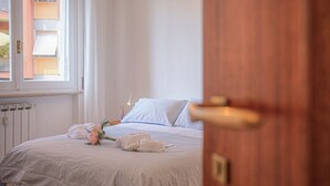 Ein ordentlich gemachtes Bett mit weißer Bettwäsche und einem Blumenstrauß, eine Holztür und ein Fenster mit Jalousien.