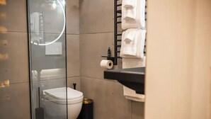 Une salle de bain moderne avec une douche à parois de verre, des toilettes blanches, une poubelle noire et un porte-serviettes avec des serviettes pliées.