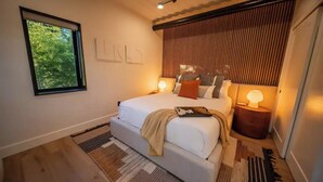 Une chambre moderne avec un grand lit, des lampes de chevet, une fenêtre donnant sur la forêt et une tête de lit en lattes de bois.