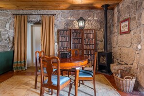 Um quarto acolhedor com parede de pedra, mesa de madeira com cadeiras, estante de livros, lareira e um sofá verde.