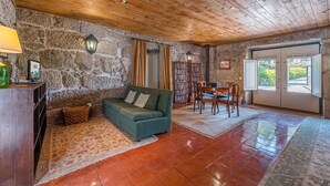 Ein gemütlicher Raum mit Steinwand, Holzboden, einem grünen Sofa, einem Holzesstisch mit Stühlen, einem Bücherregal und einem großen Fenster.