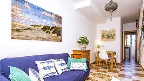 Ein Wohnzimmer mit blauem Sofa, einem Esstisch mit Stühlen, einem Schachbrettboden und einem Gemälde, das eine Strandansicht zeigt.