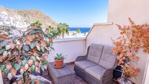 Ein Balkon mit einer Rattan-Couch, Blumentöpfen und Blick auf das Meer und Gebäude.