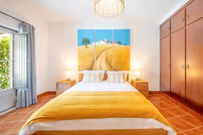 Ein Schlafzimmer mit einem Bett, zwei Nachttischen, einem großen Fenster mit Vorhängen, einem Wandbild und einem Kleiderschrank.