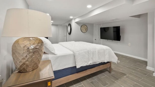 A bedroom with a bed, a nightstand, a lamp, a wall clock, and a TV mounted on the wall. - The Delightful Den (Philadelphia)