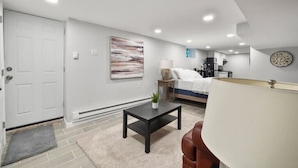 A modern, well-lit room with a bed, a small table, a lamp, and a wall clock. - The Delightful Den (Philadelphia)