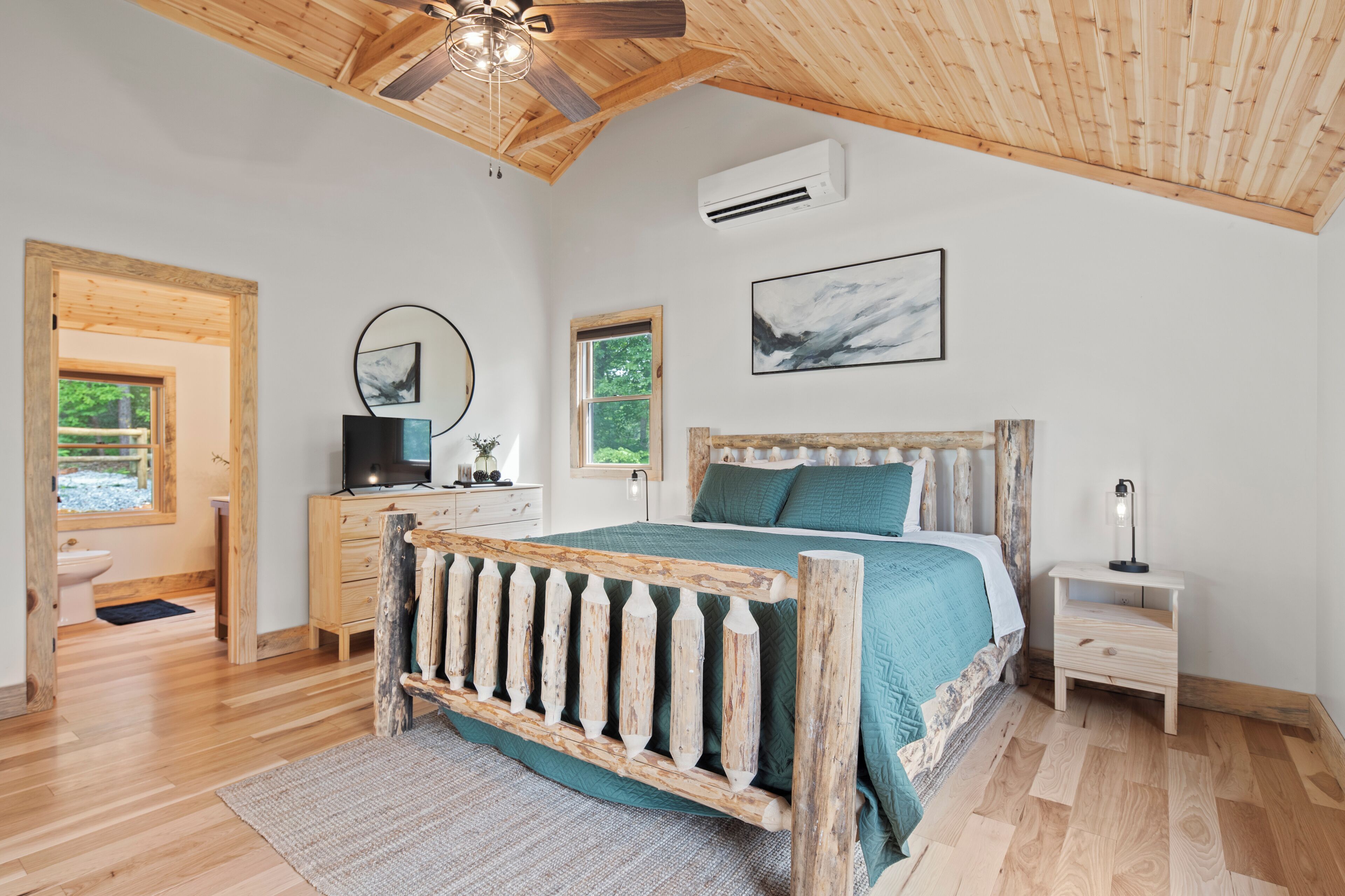 A bedroom with a wooden ceiling, a bed with a wooden headboard, a bedside table, a nightstand, and a wall-mounted air conditioner.
