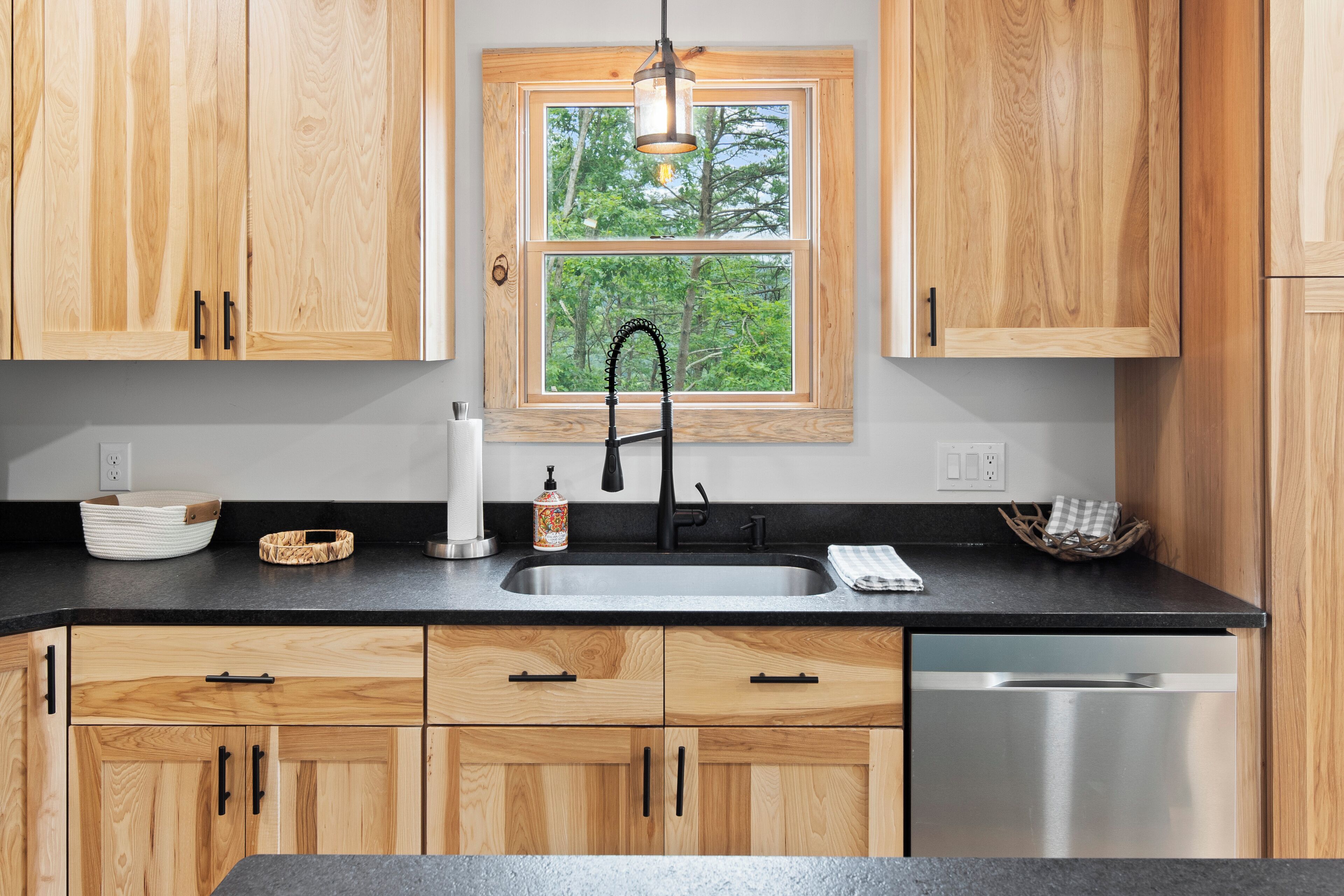 A kitchen with wooden cabinets, a black countertop, a stainless steel sink, and a window with a view of trees.