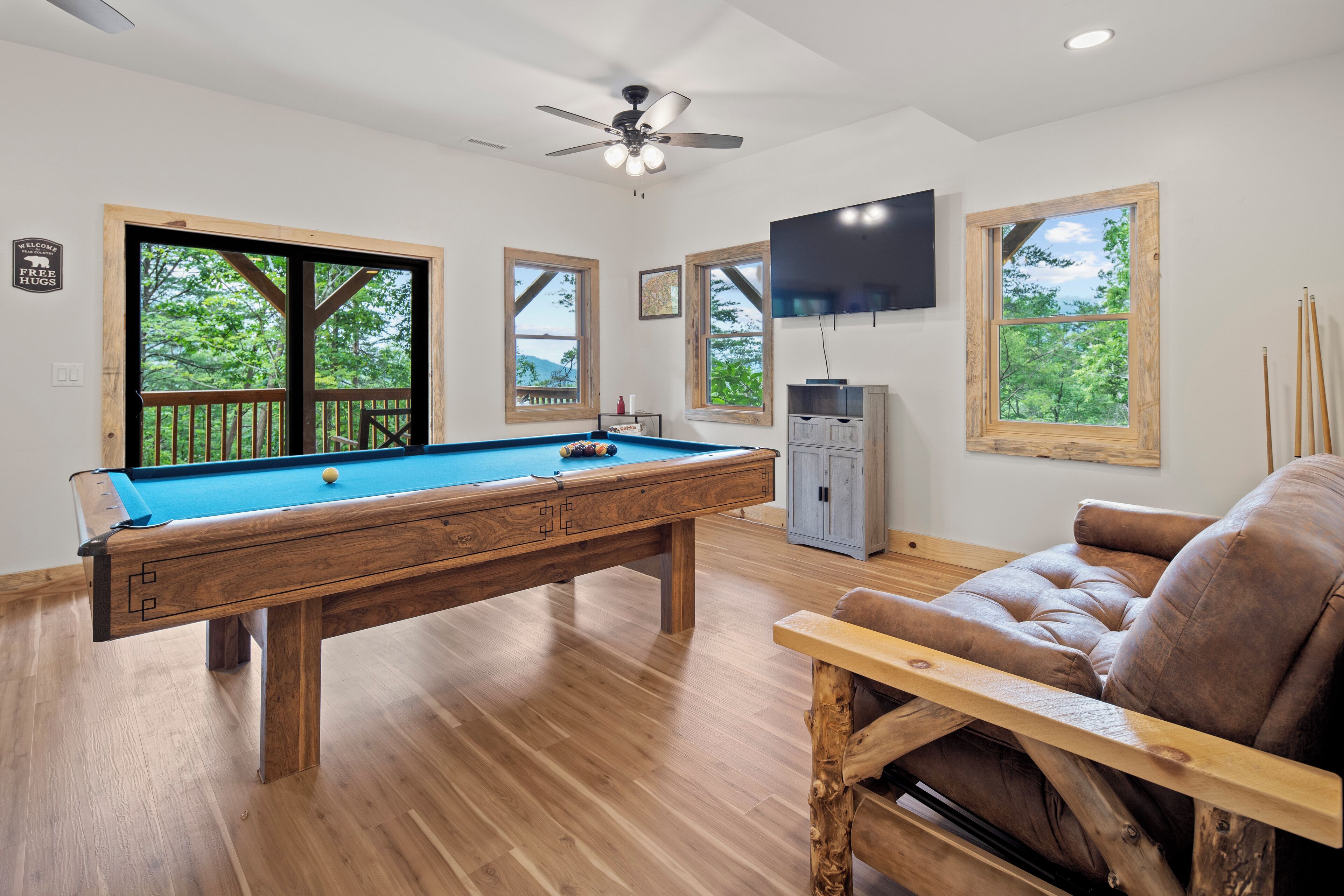 A pool table in a room with wooden flooring, a ceiling fan, and large windows with a view of greenery.