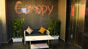 Una zona de recepción con un letrero de neón que dice "Canopy", un sofá verde con dos cojines, dos plantas en macetas y una mesa con cubierta de mármol.