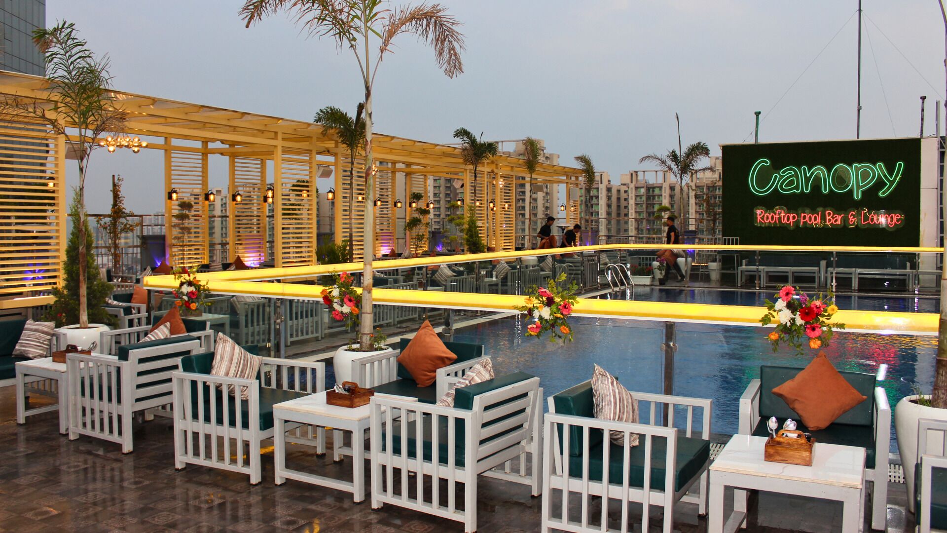 A rooftop pool area with white furniture, a neon sign for "Canopy Rooftop Pool Bar & Lounge", and a view of surrounding buildings.