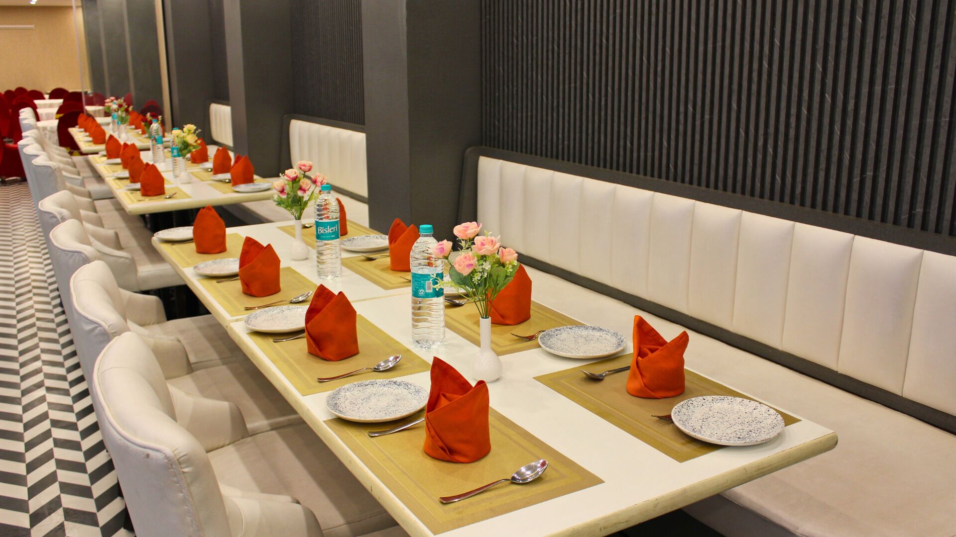 A hotel dining area with long tables set for multiple guests, each with plates, cutlery, and orange napkins.