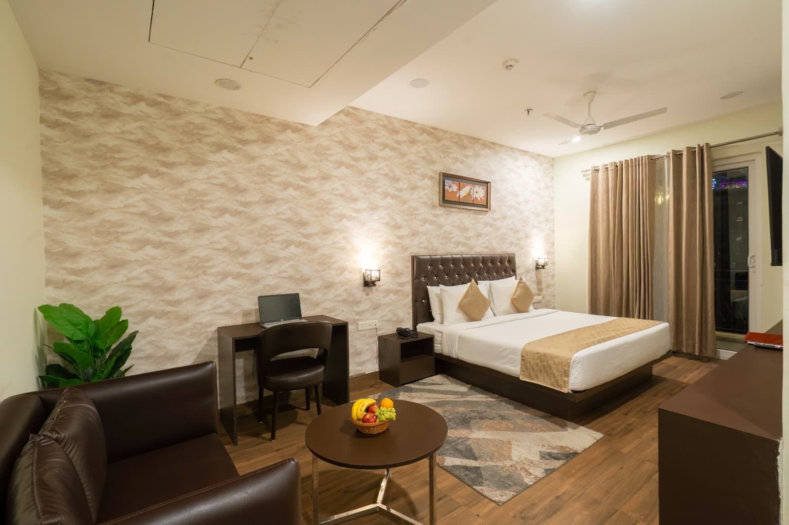 Quarto de hotel com cama, escrivaninha, cadeira, mesinha com fruta e uma planta.