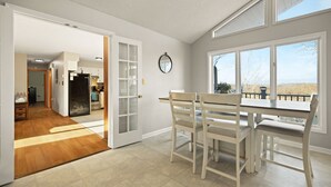A dining area with a table and chairs, a kitchen in the background, and a view through large windows.