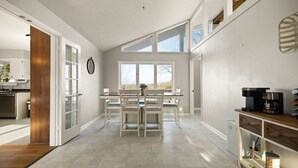 A modern kitchen with white cabinetry, a dining area with a wooden table and chairs, and large windows providing natural light.