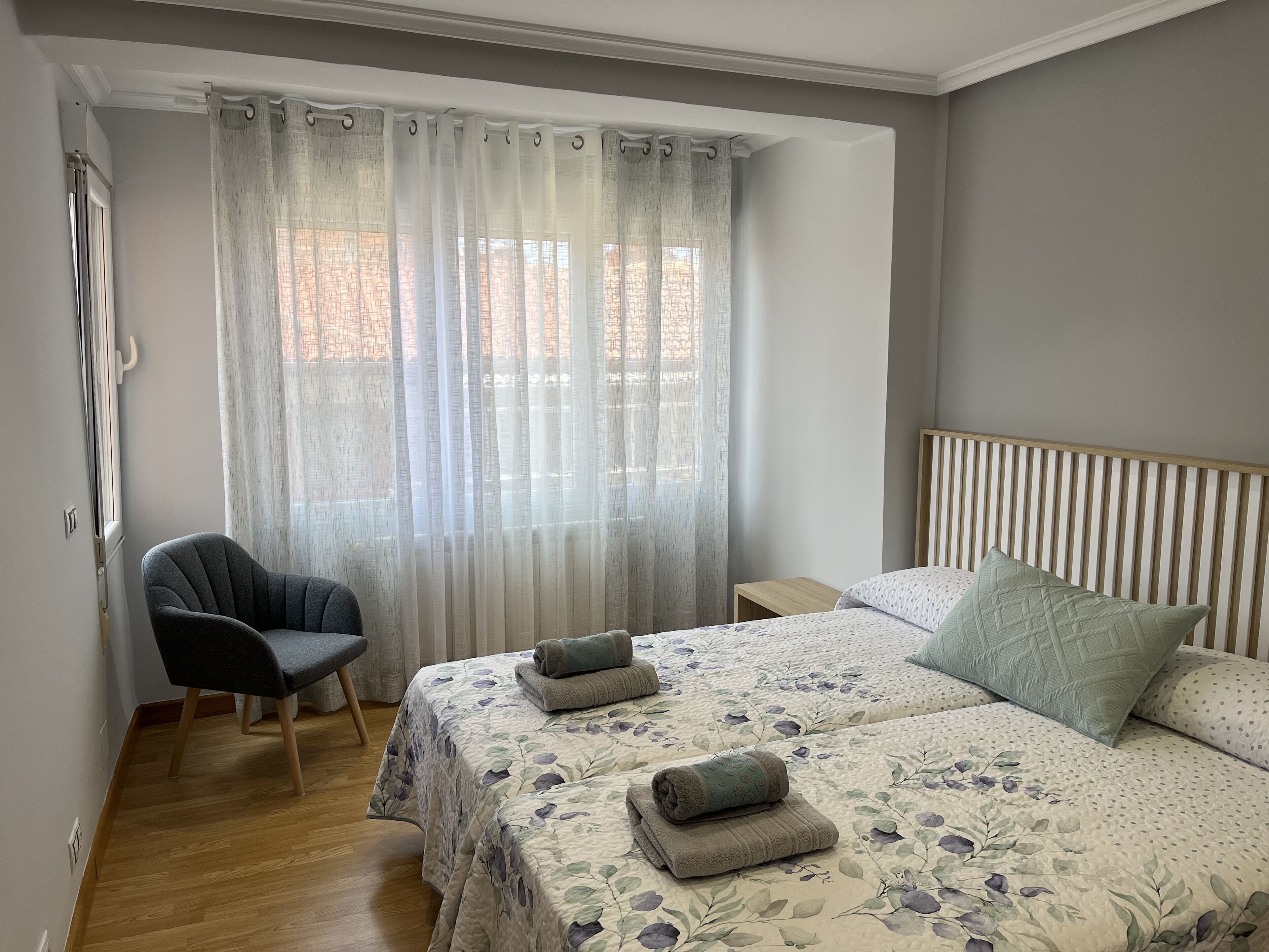 Un dormitorio con una cama, dos mesitas de noche, una silla y una ventana con cortinas translúcidas.