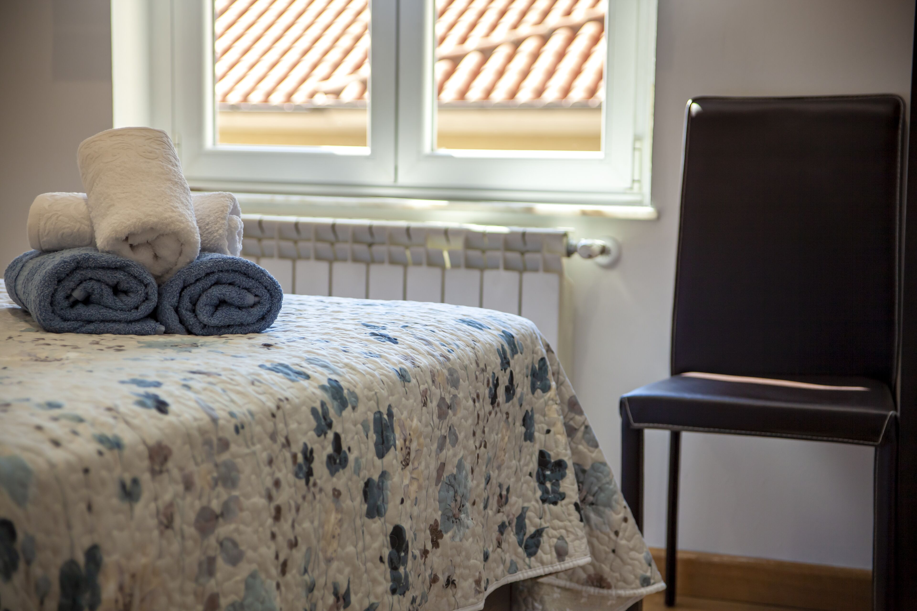 Un lit bien fait, avec des serviettes pliées, une chaise noire et une fenêtre surmontée d’un toit en tuiles.