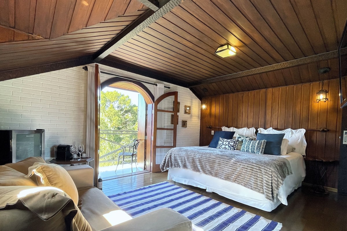 Un dormitorio con una cama grande, televisor, zona de estar con sofá y balcón con silla.