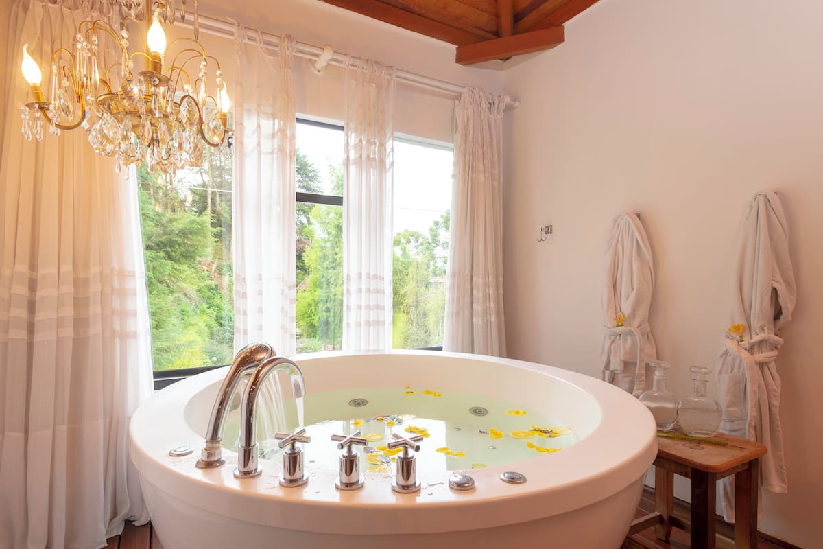 Un baño con bañera independiente, cortinas blancas, una araña y vistas al exterior con vegetación.