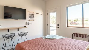 A bedroom with a bed, a TV mounted on the wall, a door leading to a balcony, and a window with a view of the outside. - The Fremont - Studio Tiny Home (Wayne County)