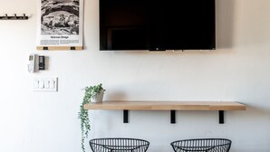 A modern living room with a mounted flat-screen TV, a framed picture of Hickman Bridge, a wall-mounted thermostat, and a wooden shelf with a potted plant and two wire chairs. - The Fremont - Studio Tiny Home (Wayne County)