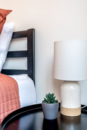 A neatly made bed with a black headboard, a white pillow, and a red blanket. A small grey potted succulent is placed on a black side table next to a white lamp with a speckled design. - The Fremont - Studio Tiny Home (Wayne County)