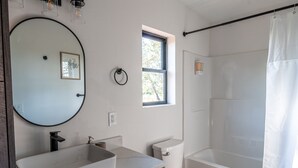 A bathroom with a large oval mirror, a rectangular sink, a toilet, and a bathtub with a shower curtain. - The Fremont - Studio Tiny Home (Wayne County)