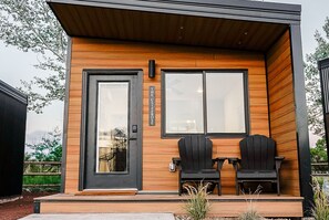 Un bâtiment moderne avec une façade en bois, deux chaises Adirondack noires sur le porche et une porte dotée d’un panneau de verre.