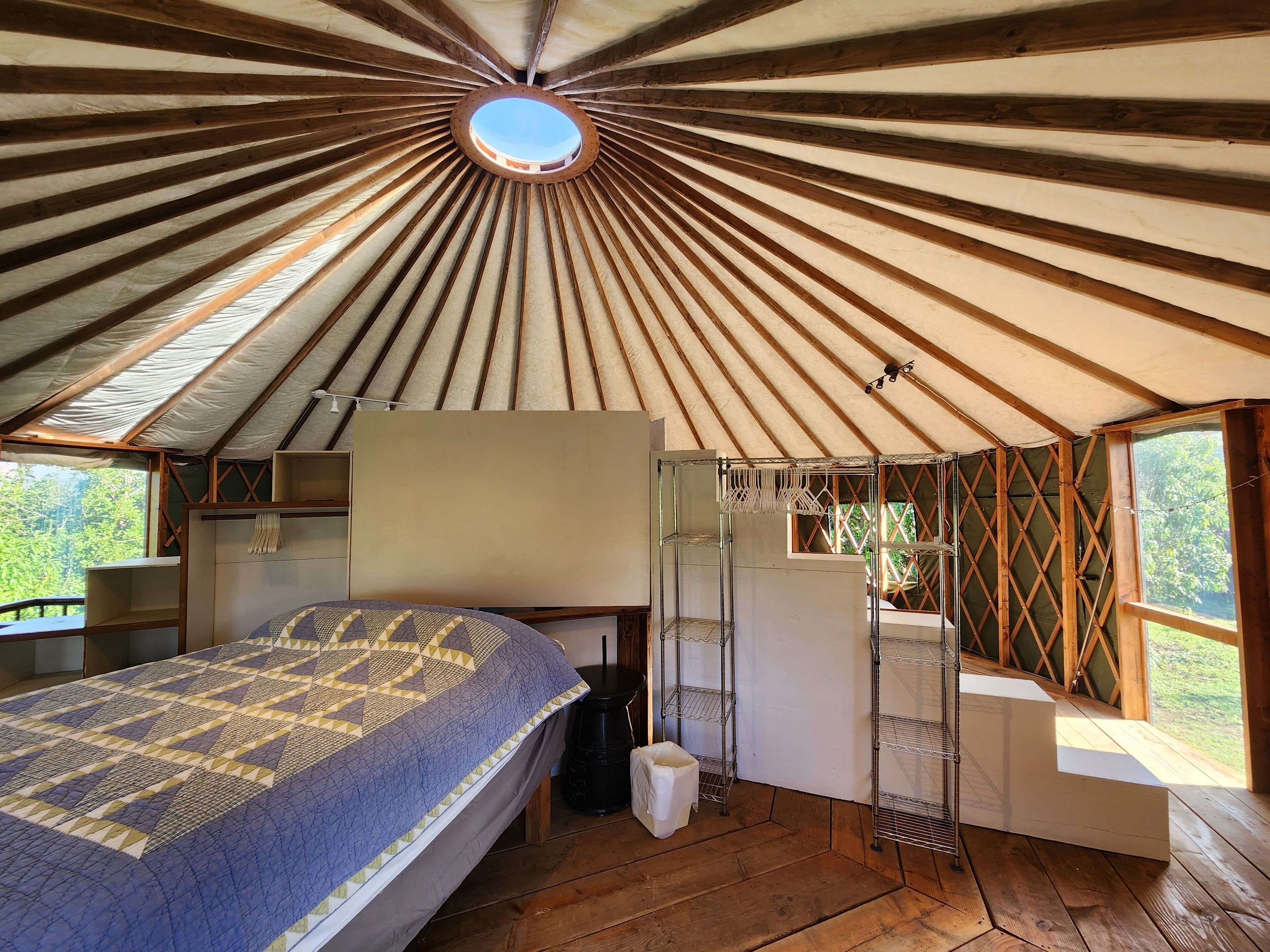 Un cuarto acogedor y circular con techo de madera y tragaluz. Se observa una cama con una colcha estampada, una mesita pequeña y una silla.