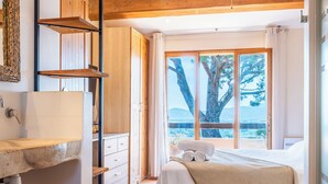 A bedroom with a bed, a wooden wardrobe, a bathroom with a sink, and a view of trees and mountains through large windows. - 5-Br. VILLA with sauna, swimming pool and sea view (La Roquette-sur-Siagne)