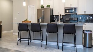 A modern kitchen with white cabinets, a stainless steel refrigerator, a microwave, and a sink. There are three black bar stools and a trash can. - Gorgeus HOUSE Near Disney & FREE water park 4963 (Kissimmee)