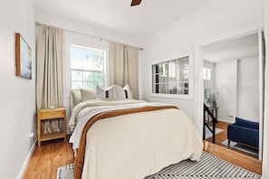 Relax in this serene bedroom with a large bed, window, and door, featuring a nightstand and lamp.