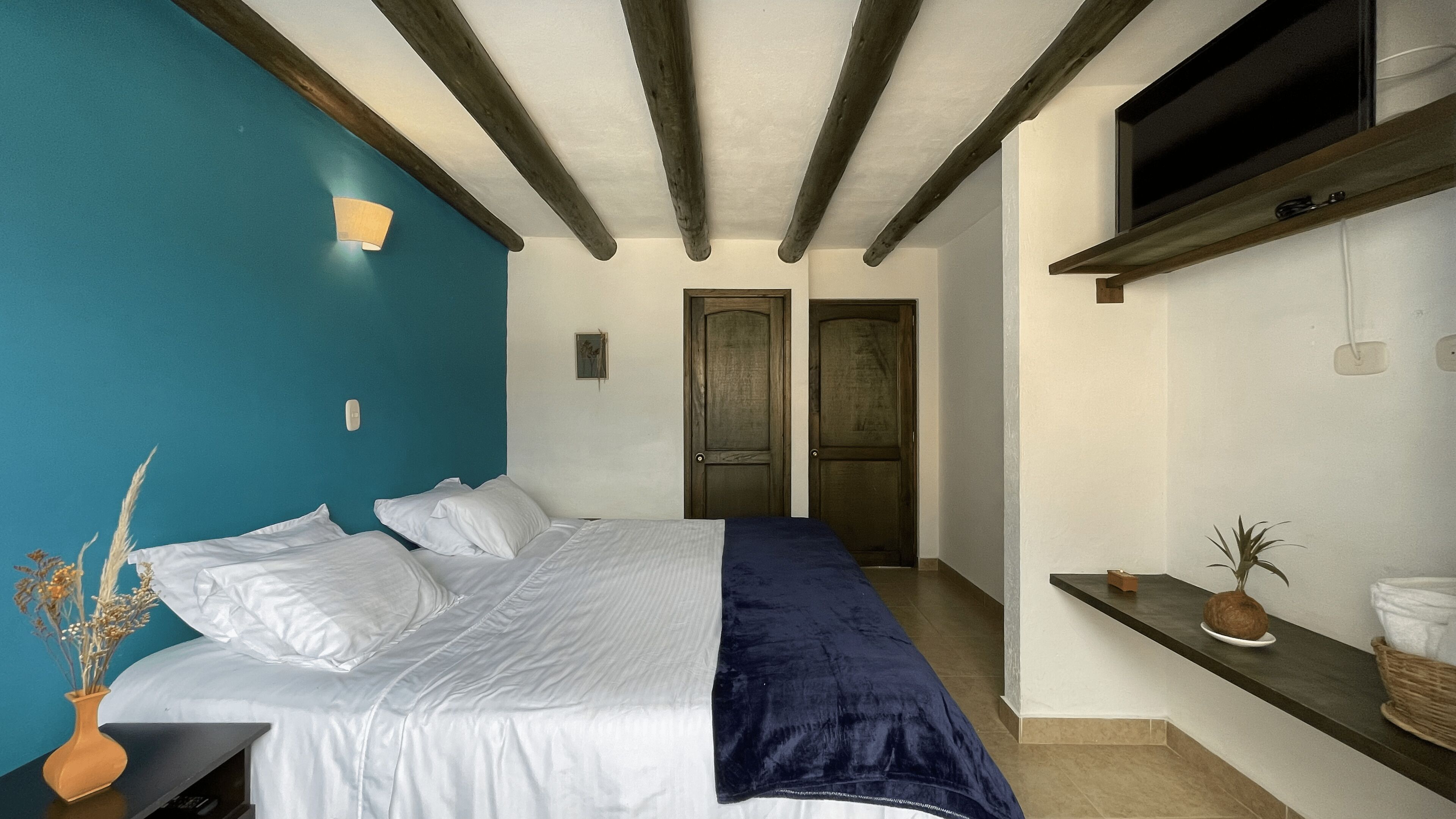 A bedroom with a bed, a blue accent wall, wooden beams, a TV, and a small plant.