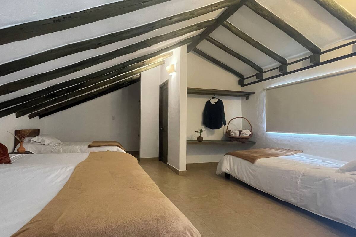 Ein Zimmer mit zwei Betten, einer Holzbalkendecke, einem kleinen Regal mit einem Korb und einer Tür zu einem weiteren Raum.