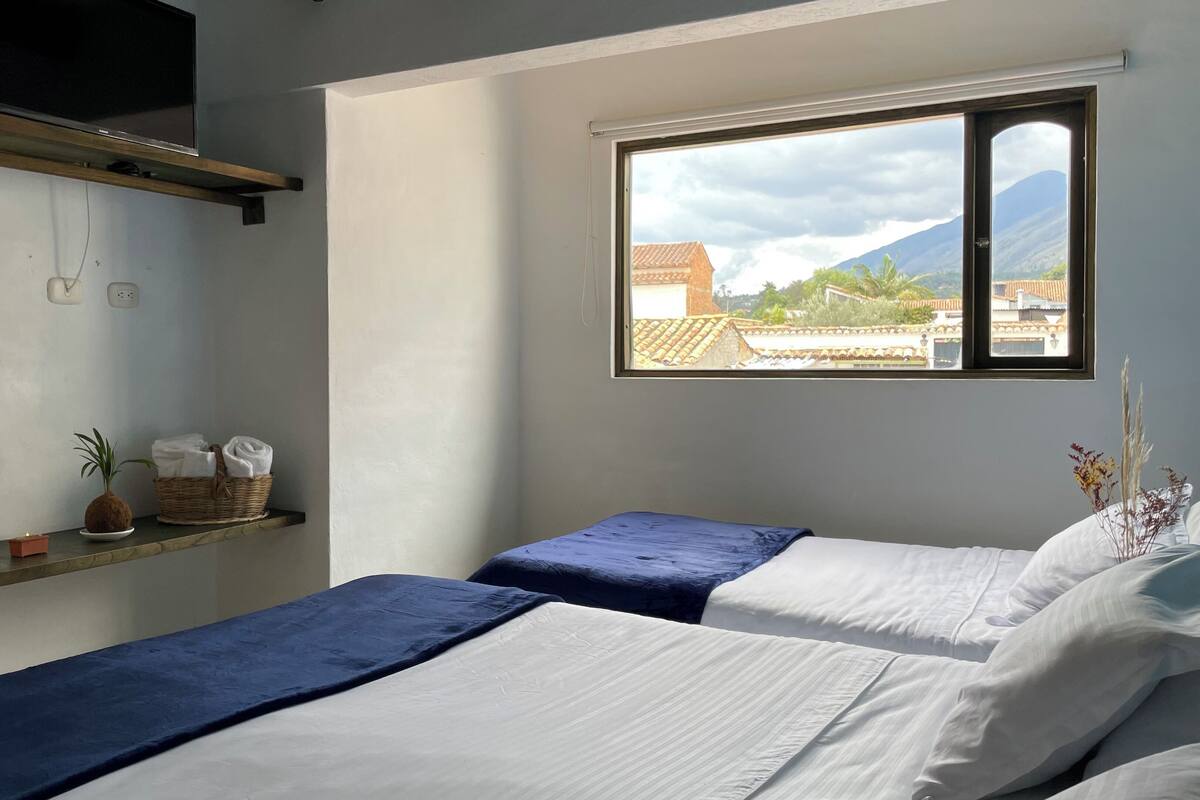 Ein Zimmer mit zwei Betten, einem Fenster mit Blick auf Gebäude und Berge, einem Regal mit Korb und Pflanze sowie einem an der Wand befestigten Fernseher.