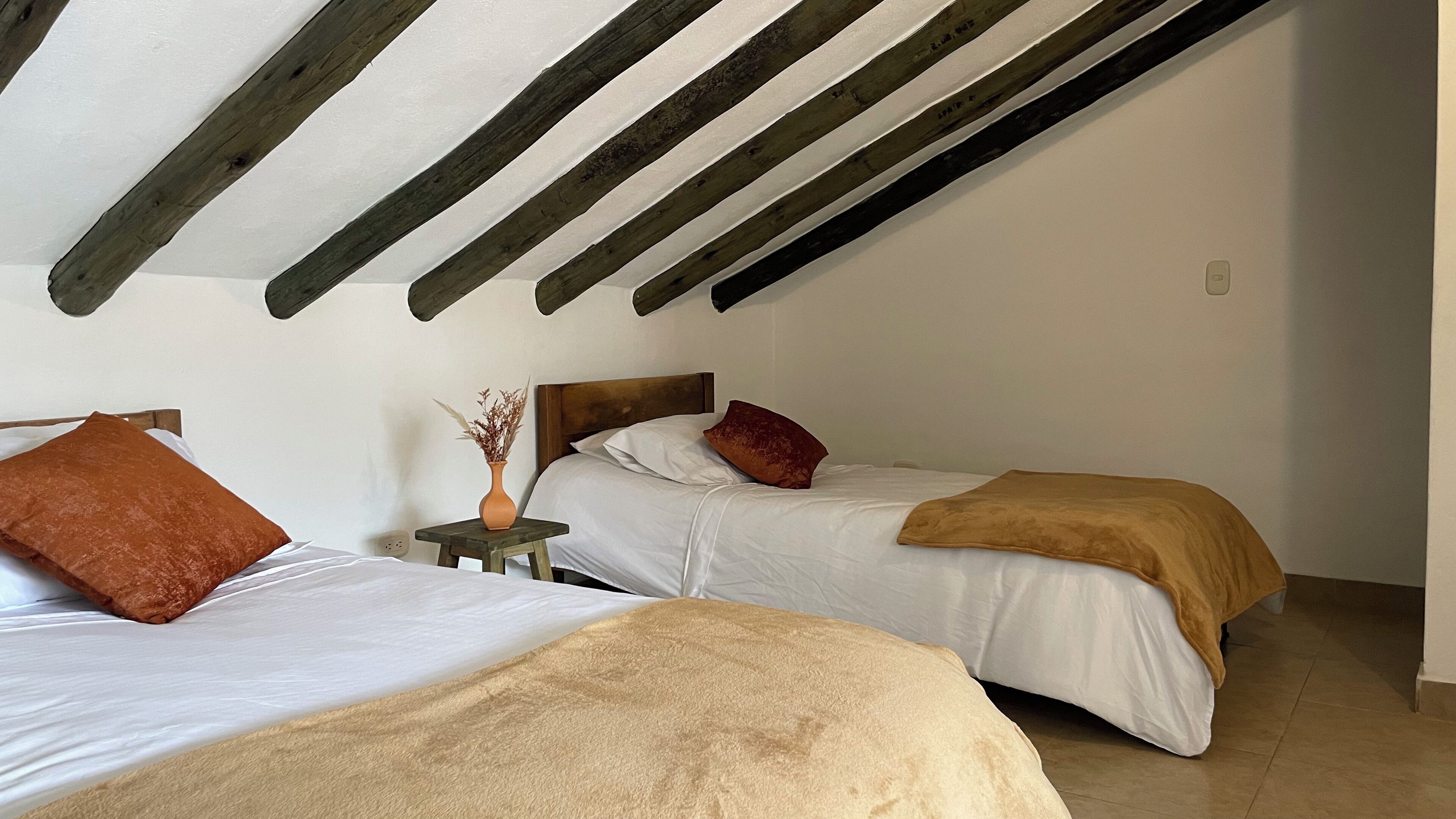 A bedroom with two beds, a small table with a vase, and a wooden ceiling beam structure.