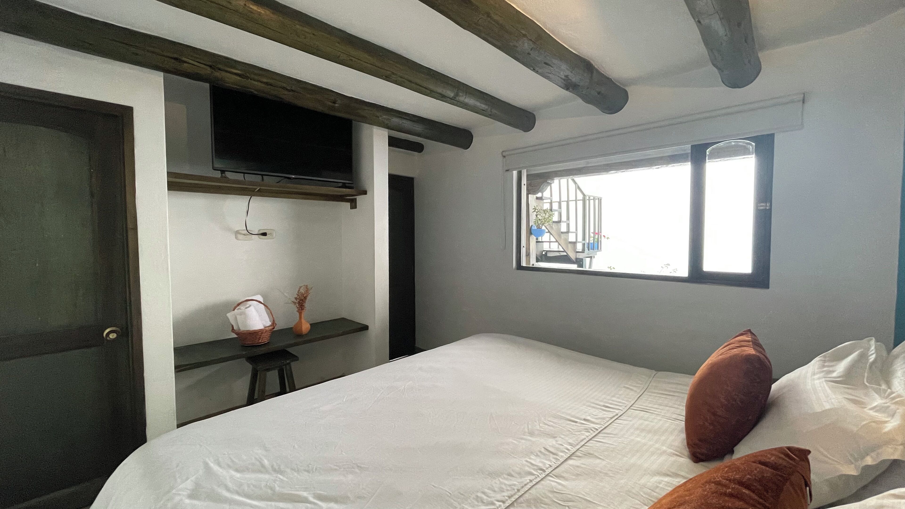 A bedroom with a bed, pillows, a small table with a basket, a vase, and a window with a view of the outside.