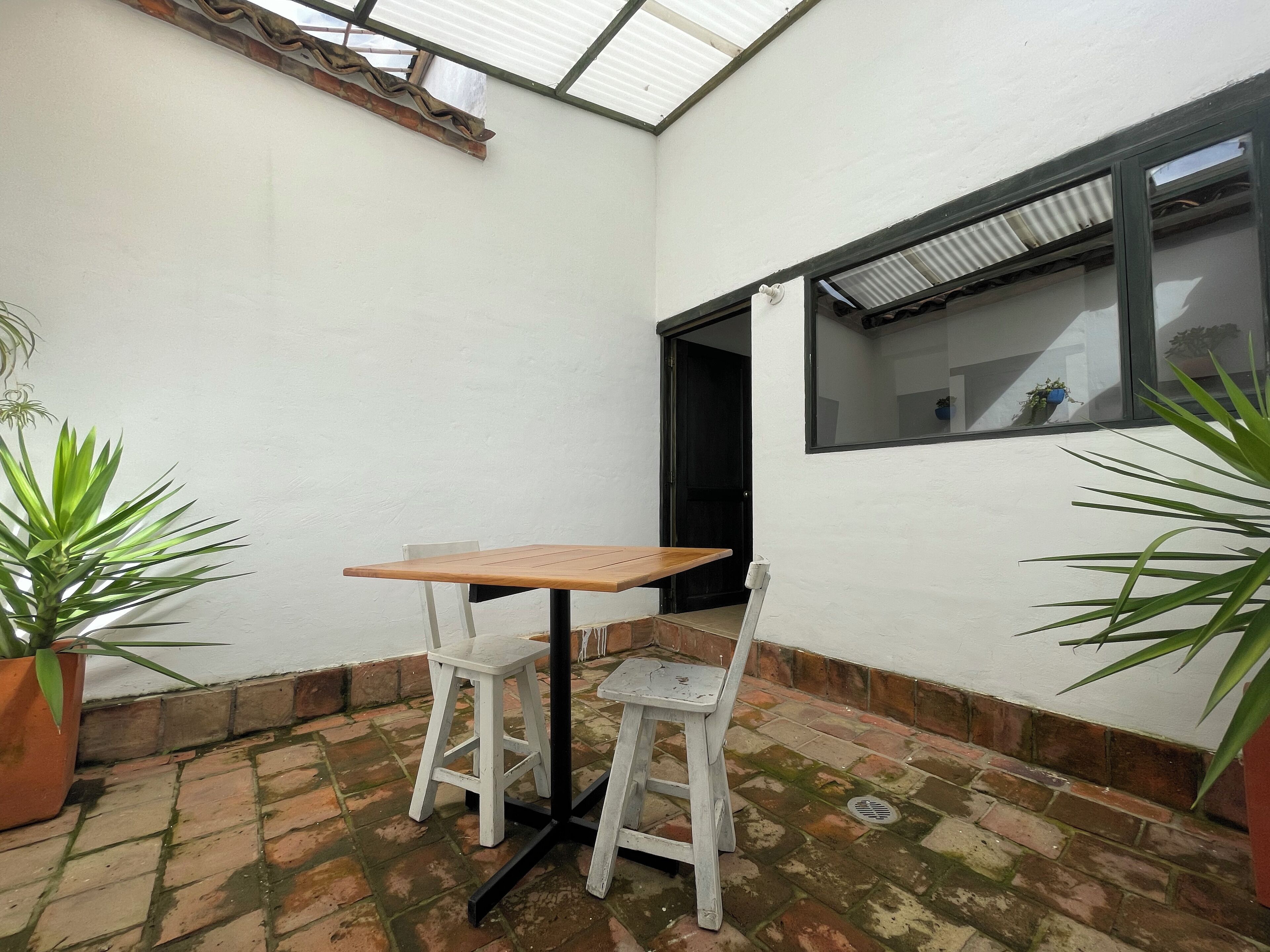 A small patio with a wooden table and two white chairs, a potted plant, and a window with a view of the interior.