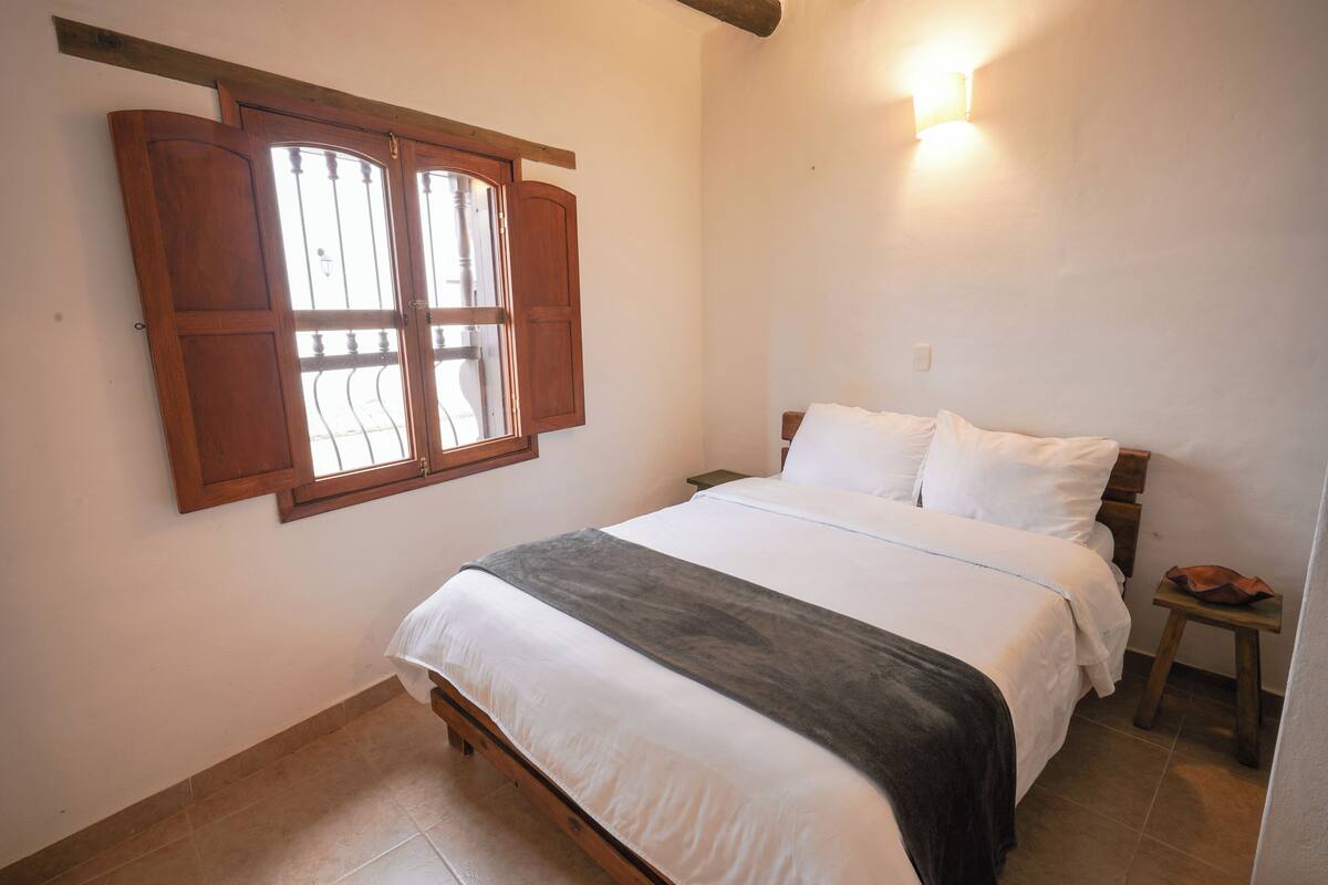 Ein Schlafzimmer mit einem Bett, einem Fenster mit Holzläden, einem Nachttisch und einer Wandleuchte.