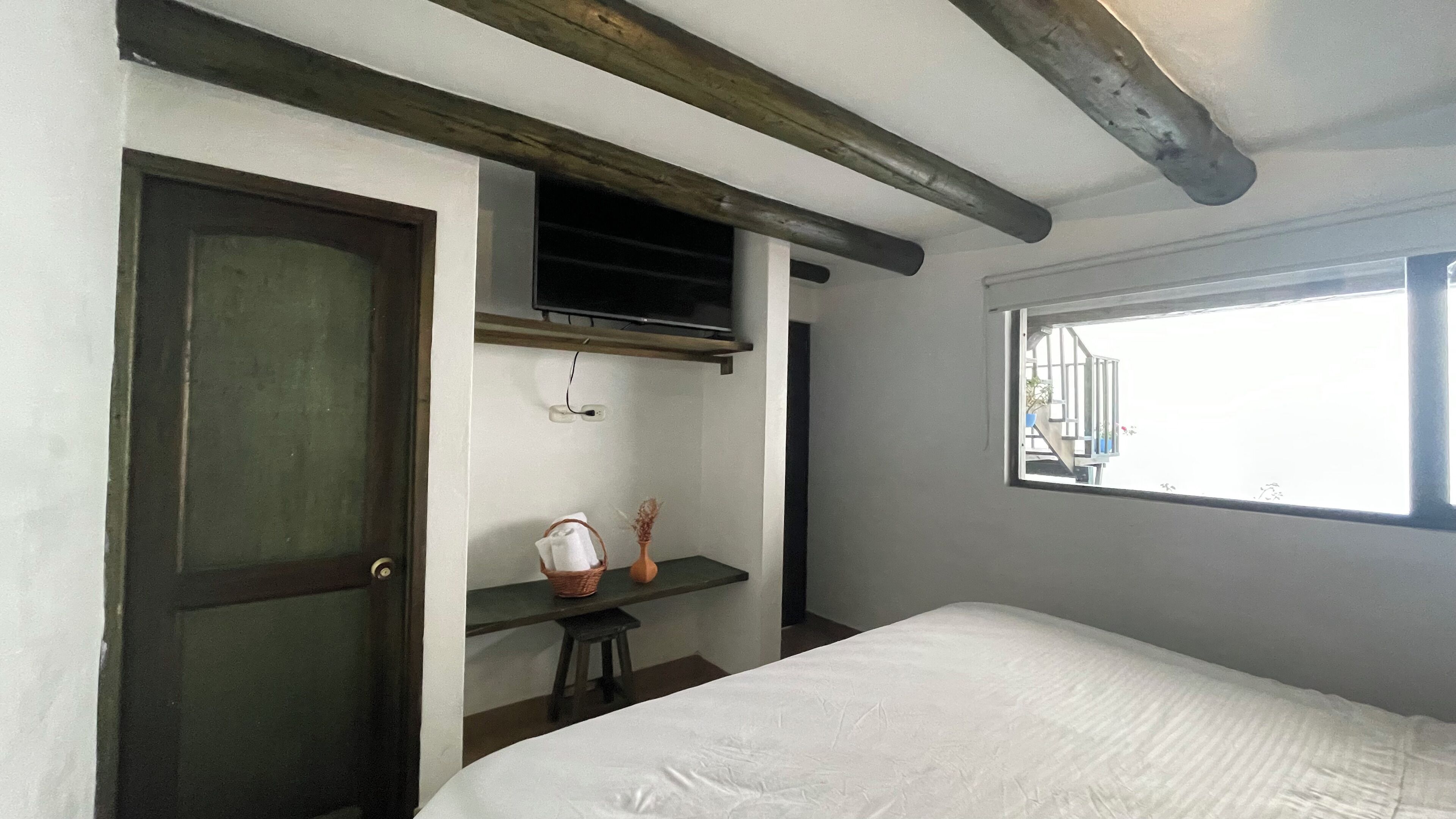 A bedroom with a bed, a window, a door, a shelf with a basket and vase, and a TV mounted on the wall.
