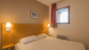 Habitación de hotel con cabecera de madera, dos lámparas de noche, un cuadro enmarcado en la pared, una ventana con cortinas rojas y un radiador.