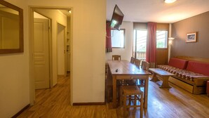 A dining area with a wooden table and chairs, a television mounted on the wall, and a sofa with red cushions.
